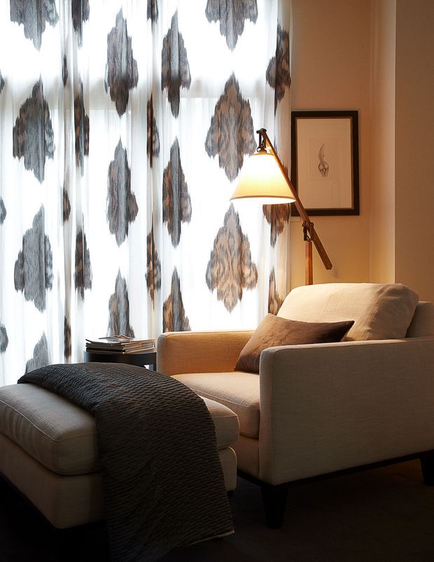 A warmly lit, compact sitting area featuring a deep upholstered armchair with matching ottoman, a dark textured throw, a timber arc floor lamp, patterned sheer curtains, and framed minimalist artwork. The space has a refined, cozy contemporary feel with neutral taupe, beige, charcoal, and warm timber tones.