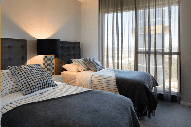 A contemporary twin bedroom with two beds dressed in white striped linens, patterned cushions, and dark charcoal throws. Upholstered charcoal headboards flank a central bedside table and black drum lamp. Full-height sheer curtains cover a large window, allowing soft daylight into the neutral, refined room.