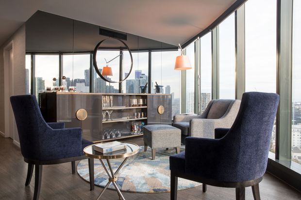 A sophisticated high-rise apartment lounge and home bar with floor-to-ceiling corner glazing overlooking a dense city skyline. Two deep navy wingback armchairs face a pair of grey upholstered lounge chairs around a small round metallic coffee table, with a patterned area rug beneath. A built-in dark timber bar cabinet with glassware and circular brass-toned door pulls anchors the room, complemented by pendant lights and a large round wall mirror.