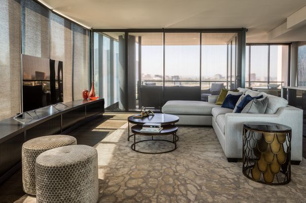 Contemporary living room with a large light-grey sectional sofa, round nested coffee tables, textured ottomans, and a low dark media console. Full-height glazing with roller blinds provides expansive city views, while patterned flooring and sculptural side tables add refined detail.