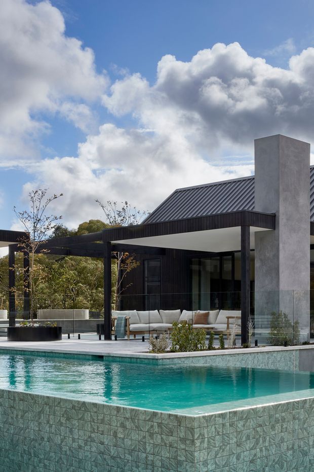 Contemporary single-storey residence with a dark metal pitched roof, black-framed glazing, a tall rendered chimney, and a covered outdoor lounge beside a turquoise tiled swimming pool. The pool terrace includes pale paving, raised planters, and minimalist outdoor furniture, set against a leafy suburban landscape beneath a dramatic blue sky.