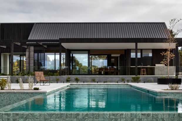 Contemporary single-storey residence viewed from the pool, featuring a long dark timber-clad facade, broad floor-to-ceiling glazing, a prominent pitched metal roof, and a covered outdoor terrace overlooking a geometric swimming pool. The restrained landscaping and concrete paving create a refined, modern outdoor setting.