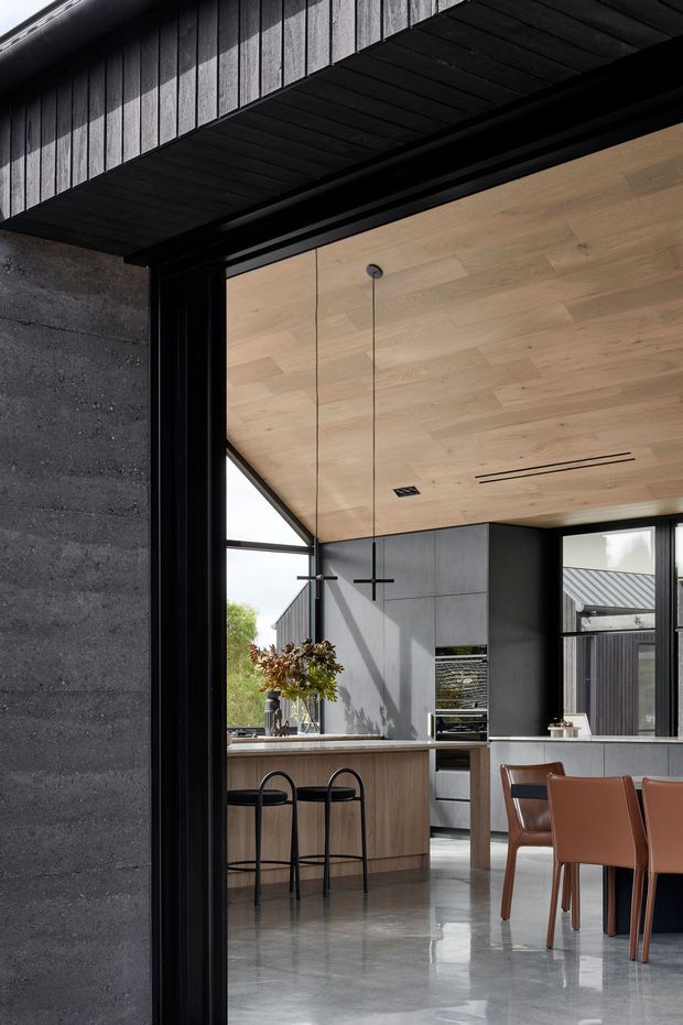 A sophisticated open-plan kitchen and dining area viewed through a large black-framed opening. The space features a warm timber-lined ceiling, dark grey wall panels and cabinetry, a central island with timber cabinetry and seating, polished concrete flooring, slim black pendant lights, and a contemporary dining table with leather chairs. Exterior cladding and roof forms are visible beyond the glazing, reinforcing the building’s dark, modern architectural character.