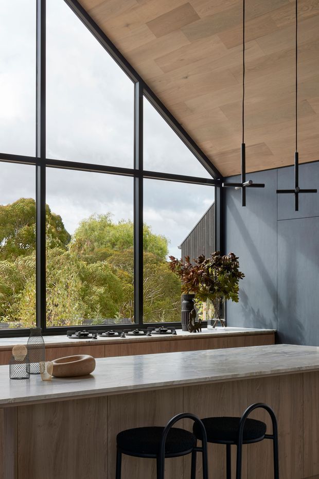 A refined contemporary kitchen with a large central island, pale stone benchtop, timber-look cabinetry, black metal-framed floor-to-ceiling glazing, and a timber-lined sloping ceiling. Two black pendant lights hang above the island, while a gas cooktop sits along the window wall. Trees are visible outside through the expansive glazing.