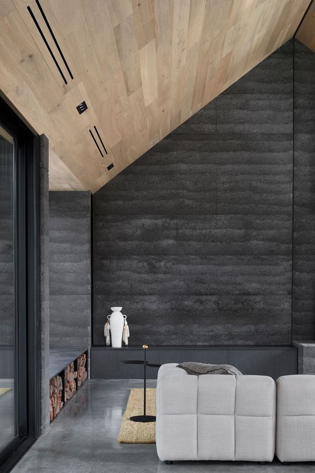 A moody contemporary living room with a dramatic dark textured feature wall, charcoal polished concrete flooring, and a warm pale timber-lined sloping ceiling. A light grey modular sofa faces the feature wall, with a slim black side table and a built-in low dark joinery unit. A black-framed glazed opening and stacked firewood are visible at the left, adding contrast and texture.