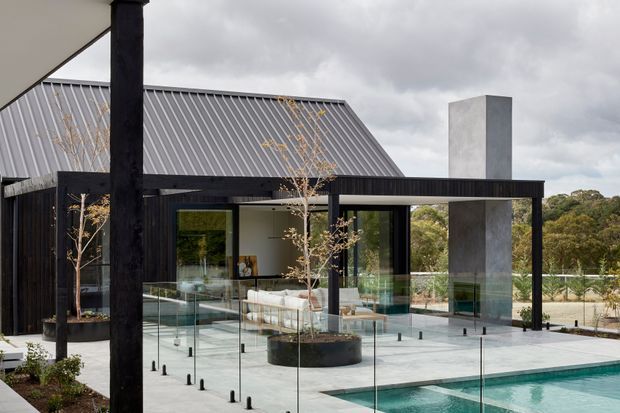 Contemporary residential exterior showing a dark-clad pavilion with broad roof overhangs, a steep corrugated metal roof behind it, expansive glazing, a tall rendered chimney, and a glass-edged swimming pool terrace. Black structural posts, pale paving, raised planter beds, and surrounding trees create a refined indoor-outdoor setting beneath an overcast sky.