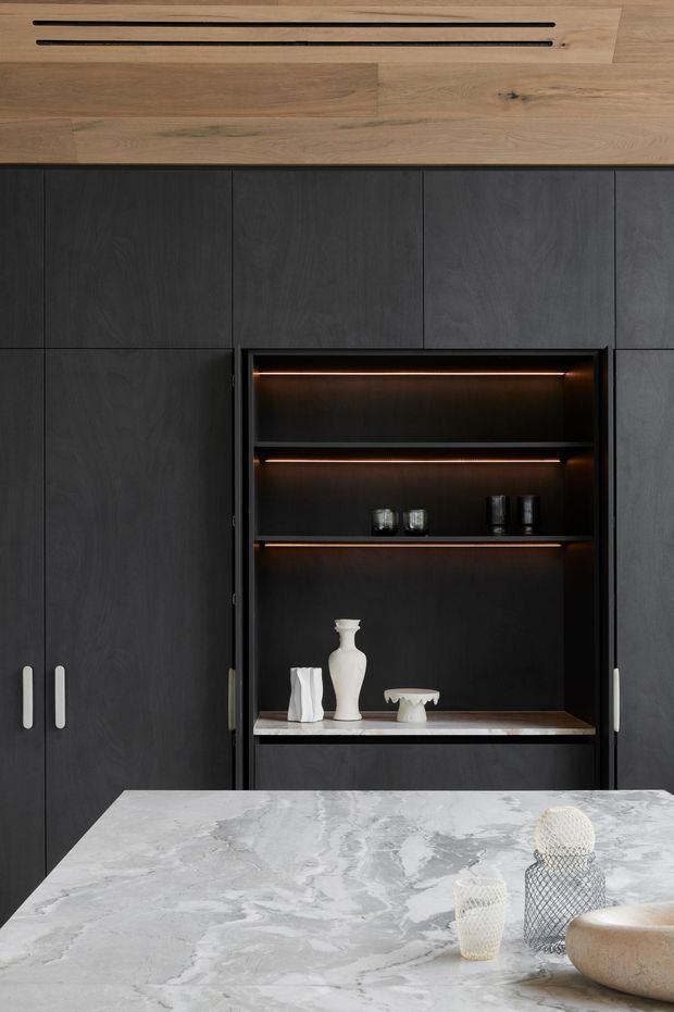 Close-up view of a sophisticated contemporary kitchen featuring floor-to-ceiling dark charcoal cabinetry with integrated vertical handles and an illuminated open display niche. Warm timber wall and ceiling detailing with linear recessed ventilation contrasts against the dark joinery. A large pale grey-and-white veined stone island benchtop dominates the foreground, with sculptural neutral-toned decorative objects.