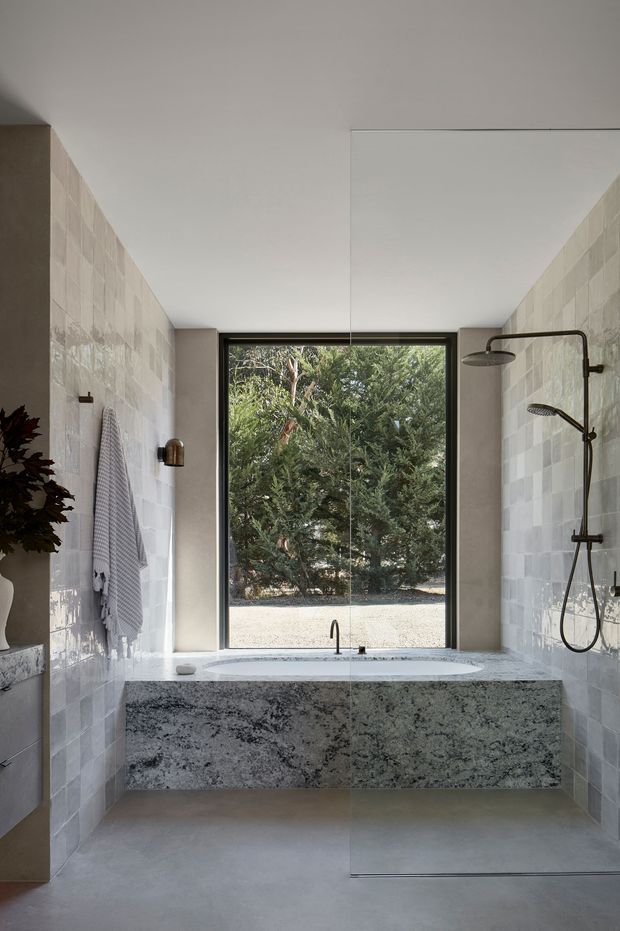 A refined contemporary bathroom with a large freestanding-style stone bathtub set into a substantial grey-speckled stone surround beneath a full-height window. Pale square wall tiles, smooth light flooring, dark-framed glazing, and a large frameless glass shower enclosure with dark metal fixtures create a calm, minimalist space. Natural greenery is visible outside.