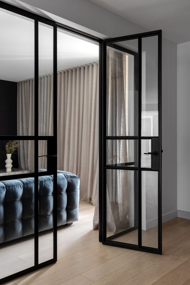 Interior view looking through black-framed glazed steel-look doors into a refined living area. The room features pale timber flooring, light grey walls, full-height neutral curtains, a deep blue tufted sofa, and a small sculptural vase. The open grid-panel door and adjacent glazed partition are the dominant architectural elements.