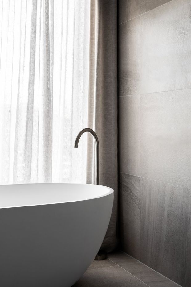 A minimalist bathroom vignette featuring a freestanding white bathtub beside a tall curved dark metal faucet. Floor-to-ceiling grey stone-look tiled walls and matching floor create a refined, monochromatic setting, softened by sheer white curtains and natural light.