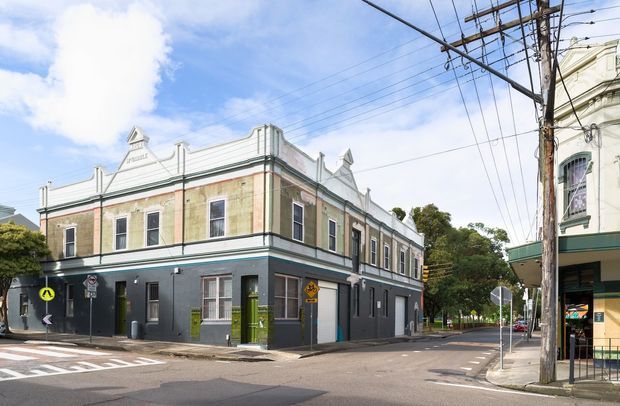 Street-level view of a heritage-style two-storey corner building with an ornate rendered facade, decorative parapet and cornice details, tall sash-style windows, and a dark painted ground floor. The building fronts a marked urban intersection with overhead utility wires and neighbouring period architecture.