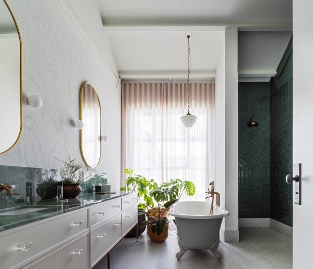 Bright, refined bathroom with a freestanding clawfoot bathtub positioned beside a large window, a long white vanity with marble or stone benchtop, green tiled splashback, arched mirrors, and a dark green tiled walk-in shower. The room combines classic fixtures with contemporary detailing and abundant natural light.