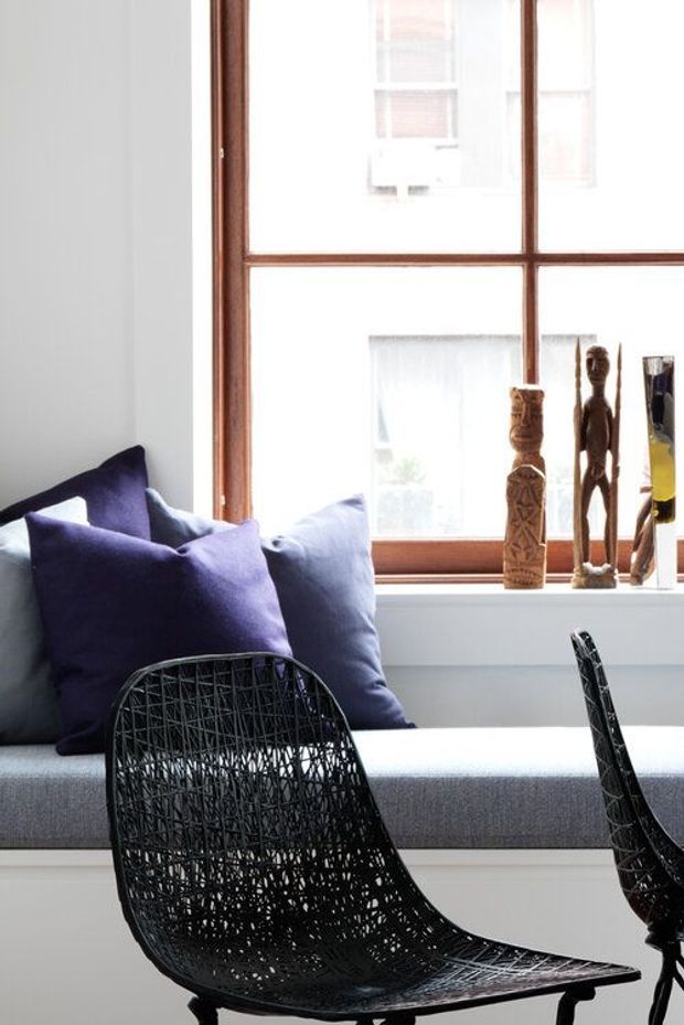 Bright contemporary interior detail featuring a built-in window seat with grey upholstery and deep purple cushions. A large timber-framed multi-pane window provides strong natural light, with carved wooden decorative objects displayed on the sill. Black woven wire lounge chairs occupy the foreground, creating a graphic contrast against the pale walls and seating.