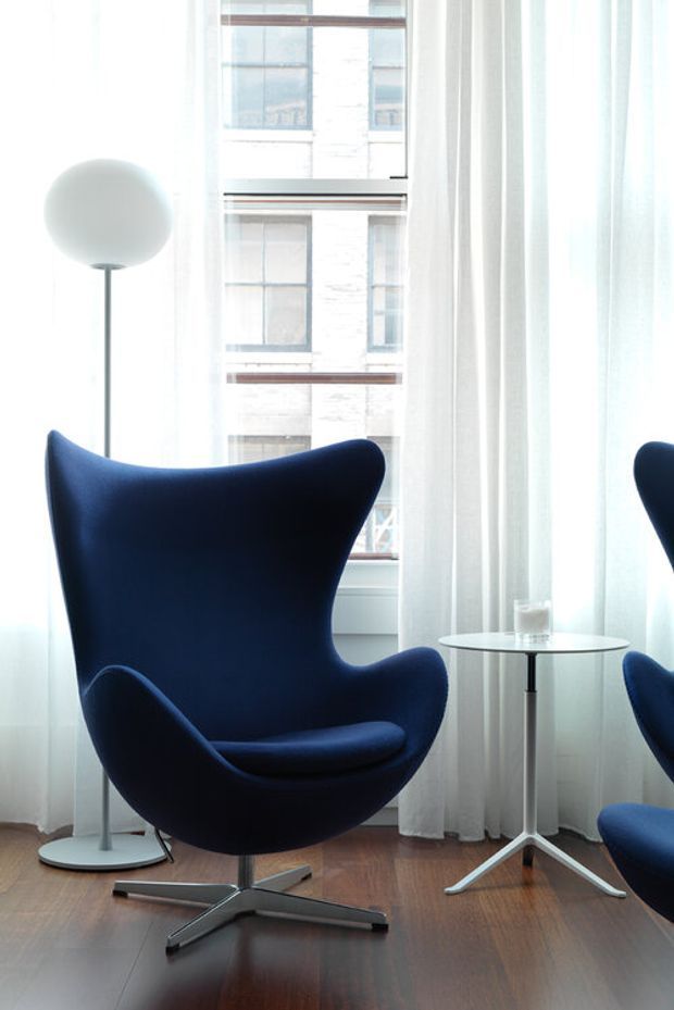 Bright, refined seating nook with a deep navy sculptural wingback lounge chair, matching partial chair, slim round side table, and spherical floor lamp. Sheer white curtains frame large windows overlooking a neighboring urban building, while dark timber flooring adds warmth.