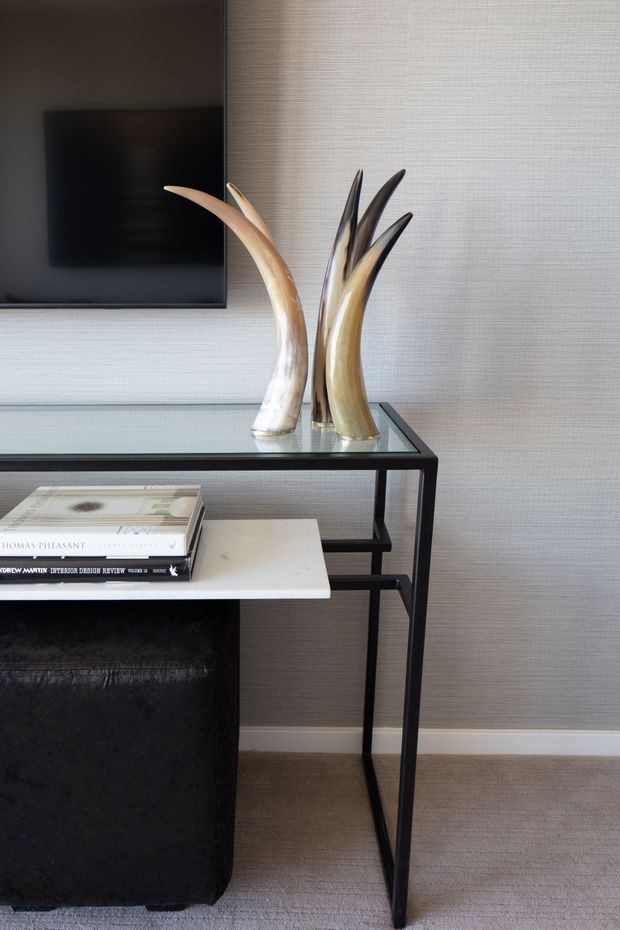A refined interior vignette featuring a slim black metal and glass console table against a textured light-grey wall. Sculptural horn-like decorative objects sit on the tabletop beside a wall-mounted black television, while stacked design books rest on a pale shelf or cabinet. A black upholstered cabinet or ottoman occupies the lower left, and beige carpet covers the floor.