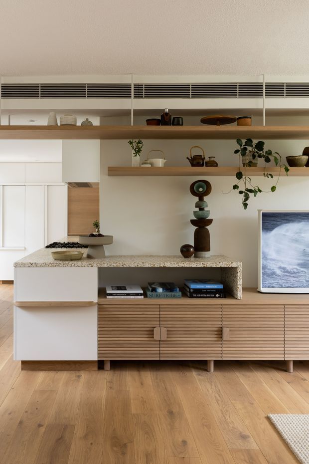 A warm, contemporary open-plan living area with a compact kitchen and built-in media cabinetry. The room features white slab-front cabinetry, a pale stone benchtop, timber flooring, and a long timber floating shelf with integrated ventilation above. A low fluted timber media console, sculptural decor, indoor plants, books, and a television create a refined, curated interior.