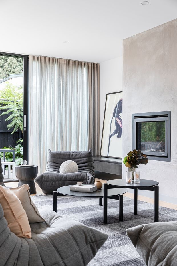A bright, contemporary living room with a layered grey sectional sofa, sculptural lounge chair, nested black coffee tables, and a textured grey rug. Full-height sheer curtains frame a large glazed opening to greenery outside. A minimalist black-framed fireplace is set into a pale textured plaster feature wall, with artwork leaning on a dark built-in ledge. The room has a clean, refined and softly neutral aesthetic.