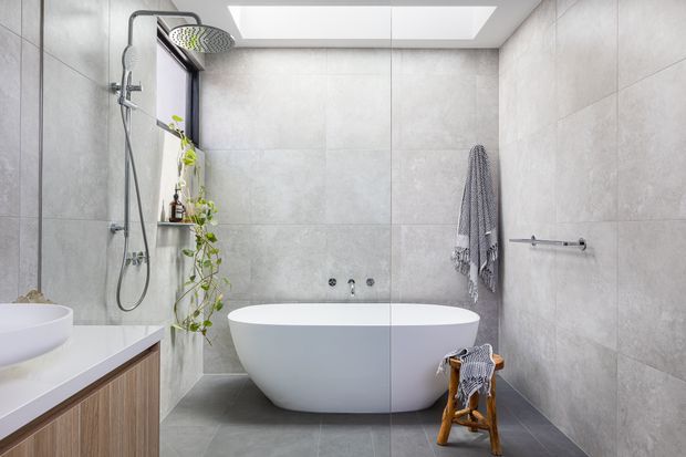 Contemporary bathroom featuring large-format grey wall and floor tiles, a freestanding white bathtub, frameless glass shower enclosure, wall-mounted rain shower, skylight, floating vanity with timber cabinetry, and trailing greenery. The space is bright, clean, and calm with a neutral grey-and-white palette.