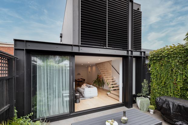 Contemporary dark-clad building exterior viewed from a private deck or courtyard. Large black-framed glazed openings reveal a warm interior living area with a timber staircase, while vertical screening and a louvred upper facade create a strong modern architectural composition. Dense greenery borders the outdoor space.