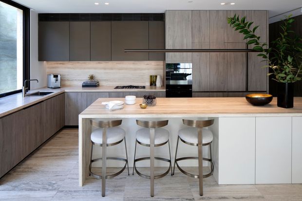 Contemporary kitchen with a large pale timber island, three stools, dark integrated cabinetry, a stone or timber-look splashback, and full-height wood-toned storage. Black-framed glazing, recessed ceiling lights, and indoor plants add contrast to the otherwise restrained neutral palette.