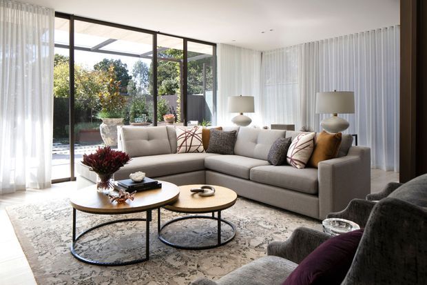 Bright contemporary living room with a large L-shaped light grey sectional sofa, two round nesting coffee tables, patterned area rug, sculptural table lamps, and sheer floor-to-ceiling curtains. Extensive black-framed glazing opens the room visually to a leafy outdoor area, creating an airy, refined interior.