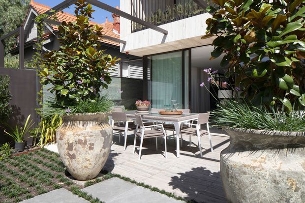 A contemporary residential courtyard and dining terrace with pale timber decking, a glass sliding door, concrete balcony and dark metal framing. A dining table and chairs sit between two large weathered stone planters filled with sculptural greenery. Neighbouring traditional red-tiled roof architecture is visible beyond the boundary, creating a contrast between the modern extension and established surroundings.