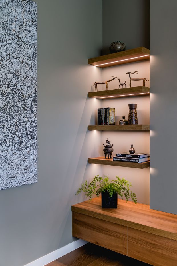 A refined built-in display and storage nook with floating timber shelves, integrated warm LED strip lighting, and a matching timber console cabinet. The shelves display books, ceramics, sculptures, and a potted fern against a grey painted wall, with a monochrome abstract artwork partially visible on the left. Dark timber flooring and clean contemporary joinery create a warm, curated interior.