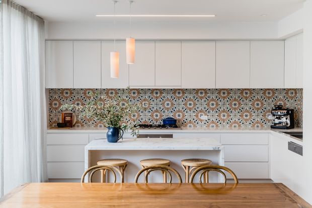 Bright contemporary kitchen and dining area with extensive white flat-panel cabinetry, a large pale stone island, timber dining table, curved timber stools, and a highly decorative patterned mosaic tile splashback. Sheer full-height curtains and simple pendant and linear lighting create an airy, refined interior.