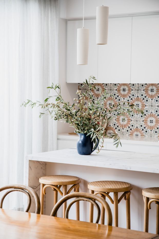 Bright, softly styled kitchen and dining area with white cabinetry, a pale marble-look island or breakfast bar, patterned decorative tile splashback, and woven rattan bar stools. Sheer curtains, pendant lights, a blue ceramic vase, and natural branches create a warm, relaxed setting. A timber dining table and curved rattan dining chairs occupy the foreground.