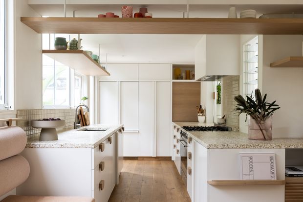 Bright contemporary kitchen with white cabinetry, timber shelving and flooring, pale stone benchtops, integrated appliances, and a central island. Open shelving displays decorative objects, while large windows provide abundant natural light. A textured tiled splashback and indoor greenery add warmth and visual interest.