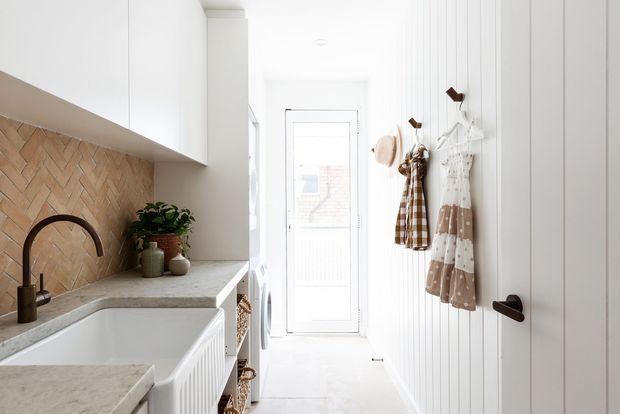 Bright, narrow contemporary kitchen and utility passage with white cabinetry, a pale stone benchtop, deep white farmhouse-style sink, warm herringbone tile splashback, dark arched tapware, and white vertical wall panelling. A glazed rear door brings in natural light, while hooks hold aprons and hats along the passage.