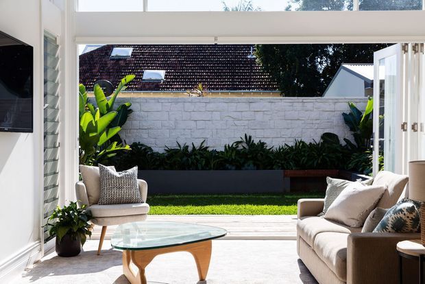 Bright contemporary living room opening through wide bifold doors onto a landscaped courtyard. The room features a cream upholstered sofa and armchair, a sculptural timber-and-glass coffee table, pale walls, and abundant natural light. Outside is a paved terrace, lawn, raised dark planter, greenery, and a rendered masonry boundary wall.