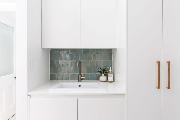 Compact minimalist white cabinetry and sink area with a muted blue-green rectangular tile splashback, integrated benchtop basin, and tall pantry-style doors with slim timber handles. A partially visible white interior door appears at left.