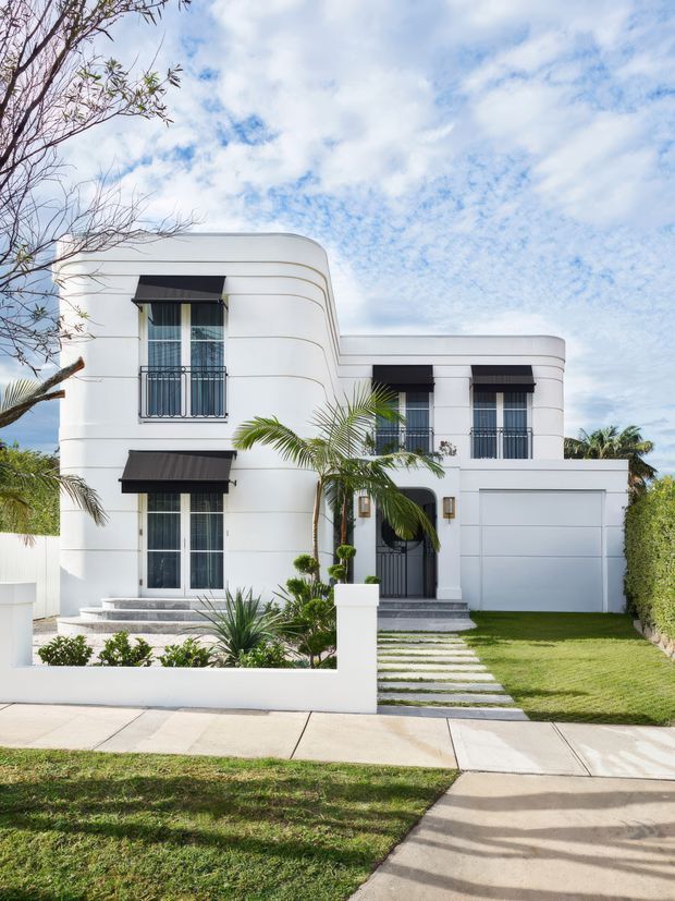 Street-facing exterior photograph of a two-storey white rendered house with rounded corners and horizontal curved detailing, black awnings, tall windows with dark metal balcony railings, a recessed entry, and an integrated double garage. A manicured lawn, palms, tropical planting, and stepping-stone path lead from the footpath to the front door.