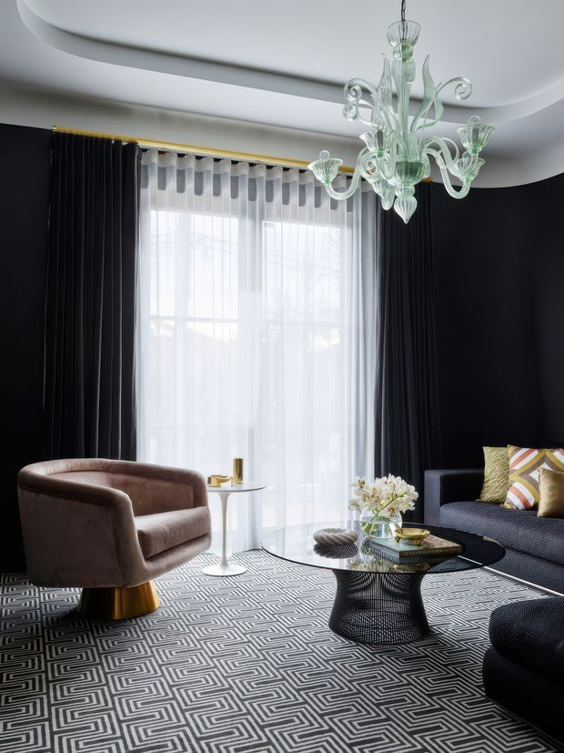 A sophisticated living room with deep charcoal walls, tall sheer-curtained glazing framed by dark drapes, and a sculptural layered ceiling. The room features a large geometric black-and-white patterned rug, a rounded taupe velvet lounge chair on a brass-toned pedestal, a dark sofa with patterned cushions, a glossy black organic-shaped coffee table, and an ornate clear glass chandelier. The overall composition is dramatic, high-contrast, and luxurious with strong Art Deco-inspired detailing.