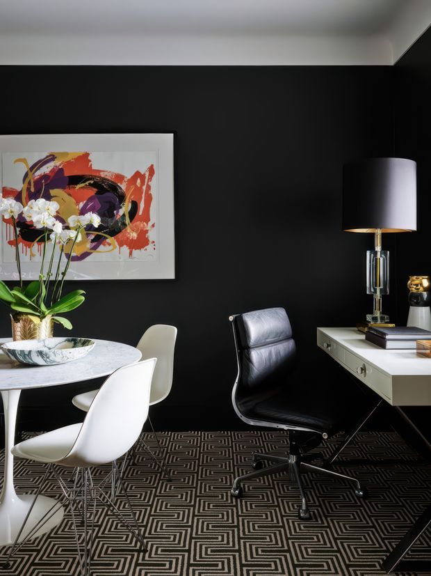 A sophisticated home office and dining area with a dramatic black feature wall, white marble round table, sculptural white dining chairs, black leather office chair, and a minimalist white desk. A colourful abstract artwork, orchid, table lamp, and geometric patterned carpet add refined decorative detail. The room has a high-contrast, glamorous contemporary feel.