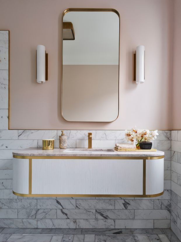 A refined bathroom vanity composition with a large rounded-rectangle mirror, flanking vertical wall sconces, a floating curved-front vanity, integrated basin and brass-toned tapware. The walls and floor are clad in veined marble, while the upper wall is painted a soft blush pink. Gold/brass trim and accessories add a polished, glamorous character.