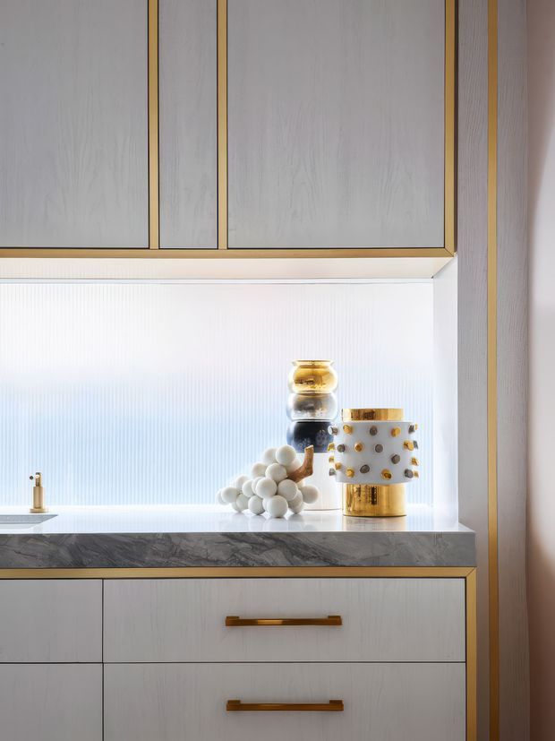 Close-up of a refined built-in kitchen or bar-style cabinetry composition, featuring pale vertical-grain cabinetry with warm brass-gold trim and handles, a dark veined stone benchtop, a softly ribbed translucent backsplash or wall panel, and curated decorative objects including a gold-accented lamp and sculptural vessels.