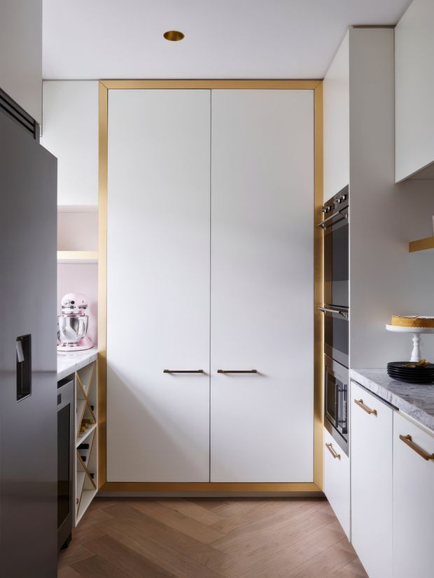 A refined, compact kitchen focused on a tall white built-in pantry or storage wall framed in brass-toned trim. White cabinetry with brass handles, integrated stainless-steel ovens, pale marble-look benchtops, a dark refrigerator, and warm herringbone timber flooring create a clean, contemporary look with subtle luxe detailing. A countertop mixer and cake stand are visible at the sides.