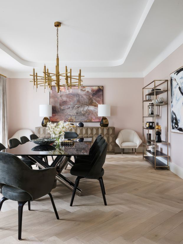 A refined dining room with a dark glass dining table surrounded by black upholstered chairs, set against pale beige walls and light herringbone timber flooring. A sculptural brass chandelier hangs above the table, while a geometric patterned sideboard, abstract artwork, lamps, open shelving, and upholstered occasional chairs create a polished contemporary interior.