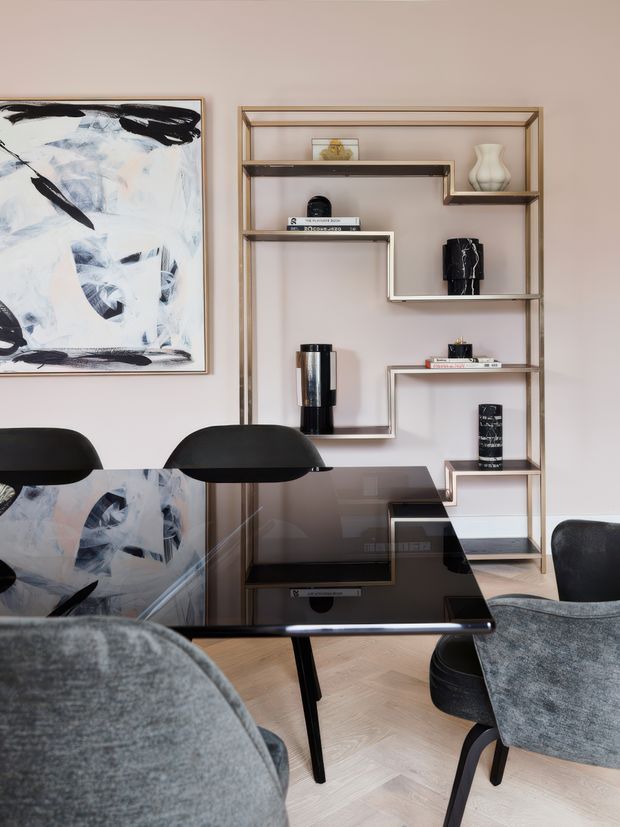 A refined dining area with a glossy black rectangular dining table and upholstered grey dining chairs. A large abstract artwork hangs on a soft neutral wall beside an open geometric shelving unit in a metallic frame, styled with books, ceramics and dark decorative objects. The room has pale herringbone timber flooring and a restrained neutral palette with black accents.
