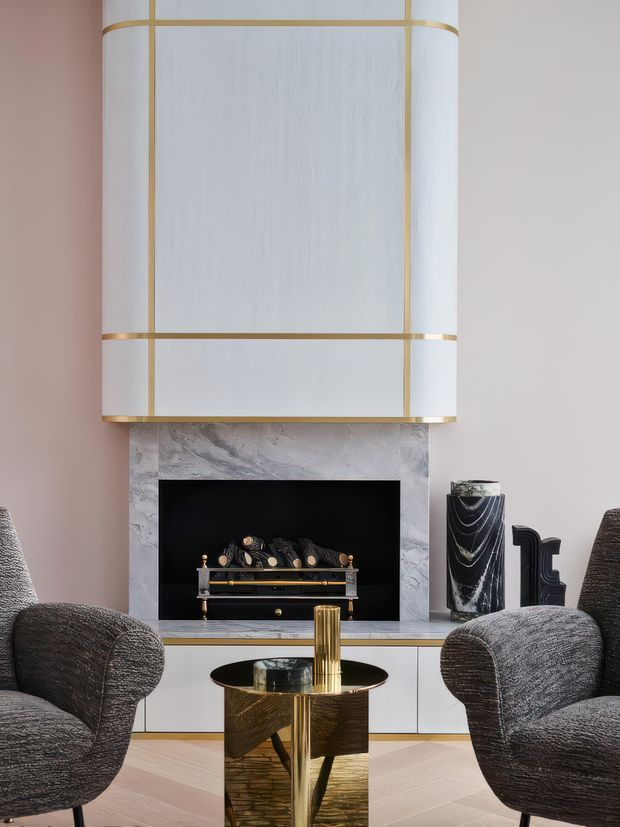 A refined living room centred on a sculptural fireplace with pale veined stone surround, black firebox and brass detailing. Two textured dark-grey lounge chairs face a small reflective brass-and-glass coffee table, while a large cylindrical white pendant with brass bands hangs prominently above. Pale timber flooring, soft blush walls, monochrome accessories and metallic accents create a polished, high-contrast interior.
