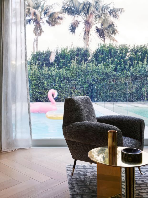 A stylish indoor lounge area beside full-height glazing, furnished with a sculptural black armchair, round glass-topped side table, gold-toned cylindrical vase, dark bowl, and patterned rug. Pale timber herringbone flooring and sheer curtains frame a view of a swimming pool, glass balustrade, dense hedge, and palm trees outside. The interior has a refined contemporary feel with strong black, gold, and light timber contrasts.