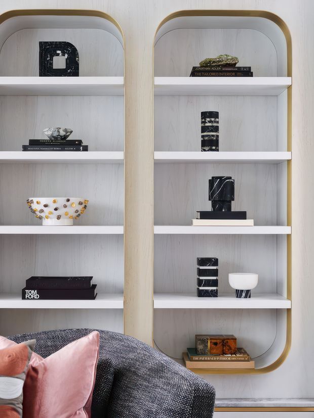 Close-up interior view of custom built-in display shelving with rounded vertical frames, pale timber-grain backing, white shelves, and subtle brass-gold edging. The shelves hold books, sculptural objects, vases, and decorative pieces. The lower foreground shows part of a dark textured sofa with blush pink cushions, creating a refined, softly styled living space.