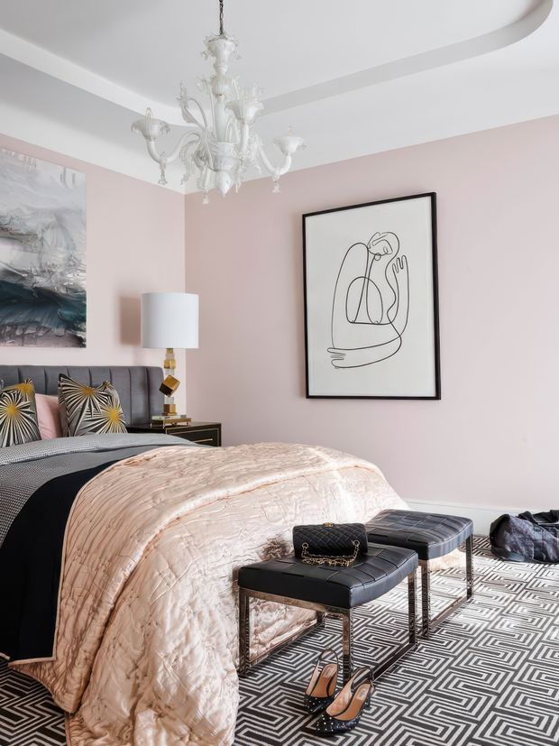 A sophisticated bedroom with blush pink walls, a white decorative tray ceiling, and an ornate white chandelier. The room features a neatly made bed with a pale blush quilt, a dark upholstered headboard, patterned accent cushions, and a black-and-white geometric rug. A black framed line-art print, bedside lamp, dark side table, and a black upholstered bench with a matching luggage-style ottoman complete the refined, glamorous interior.