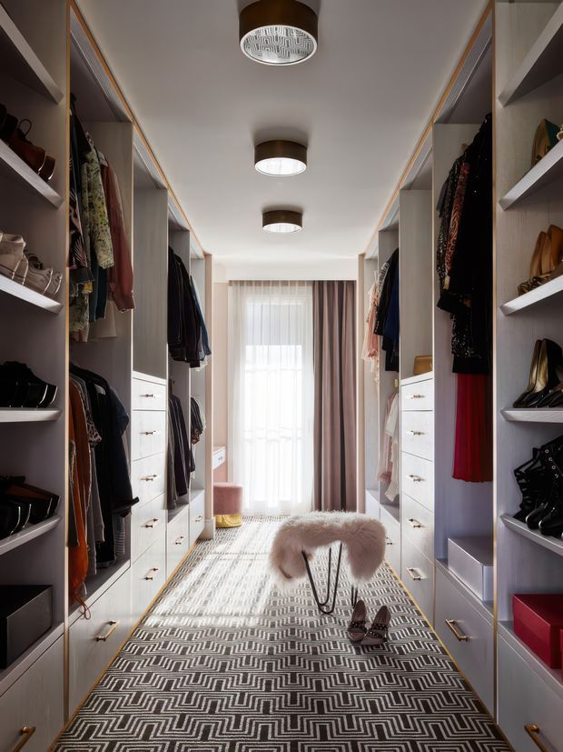 A narrow, elongated walk-in wardrobe with floor-to-ceiling open shelving, hanging rails, drawers, and shoe storage lining both sides. The space is softly lit by a series of round ceiling fixtures and receives natural light through a curtained window or glazed door at the far end. A patterned runner, small stool, and accessories create a refined dressing-room atmosphere.