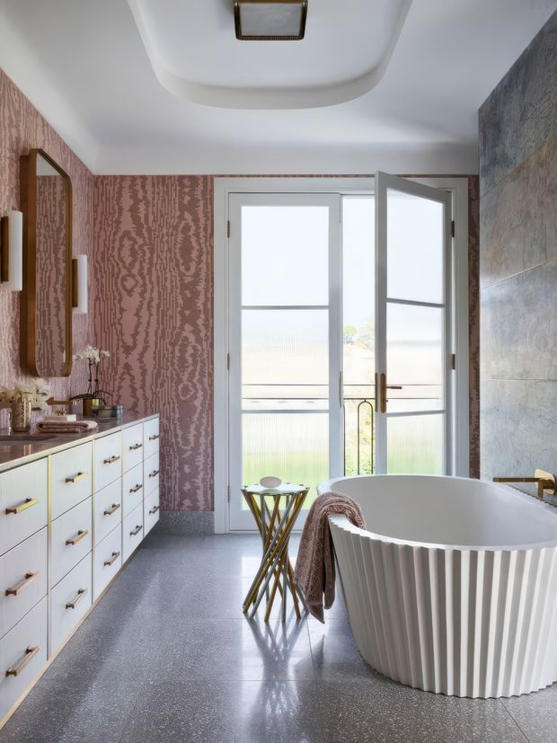 A stylish bathroom with a freestanding fluted white bathtub, large white vanity cabinetry with brass hardware, a wall-mounted mirror, patterned mauve wallpaper, and grey tiled wall surfaces. Frosted glazed double doors open to a balcony or exterior, bringing in soft natural light. The room has polished grey flooring, sculptural lighting, and refined decorative details.