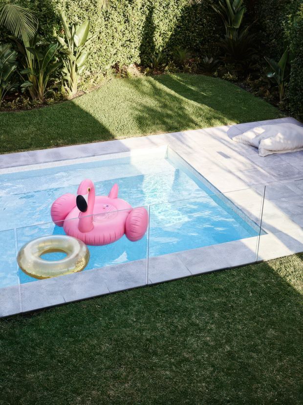 A compact outdoor swimming pool with clear blue water, surrounded by pale stone or concrete paving and transparent safety fencing. Tropical planting and manicured lawn border the pool, with a small poolside cushion and inflatable pool floats visible. The image is brightly sunlit and presents a lush landscaped backyard setting.