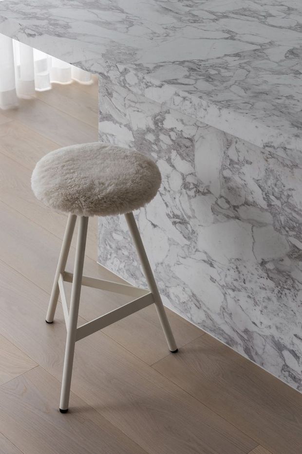 A minimalist interior detail featuring a sculptural pale upholstered bar stool with a light timber or painted frame, positioned beside a full-height veined marble wall and counter edge. The room has pale timber flooring, soft neutral tones, and curved white built-in shelving visible in the background.