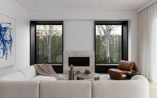 A refined contemporary living room with a large cream sectional sofa in the foreground, two black-framed windows, a marble fireplace surround, a low round coffee table, and a sculptural brown leather lounge chair. White walls, decorative ceiling moulding, sheer curtains, and a blue abstract artwork create a calm, high-end interior.